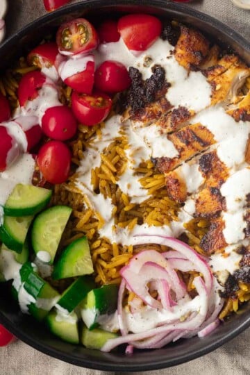 One pot shawarma and rice with grilled chicken bowl with rice, tomatoes, cucumbers, red onions, and white sauce on a black plate.