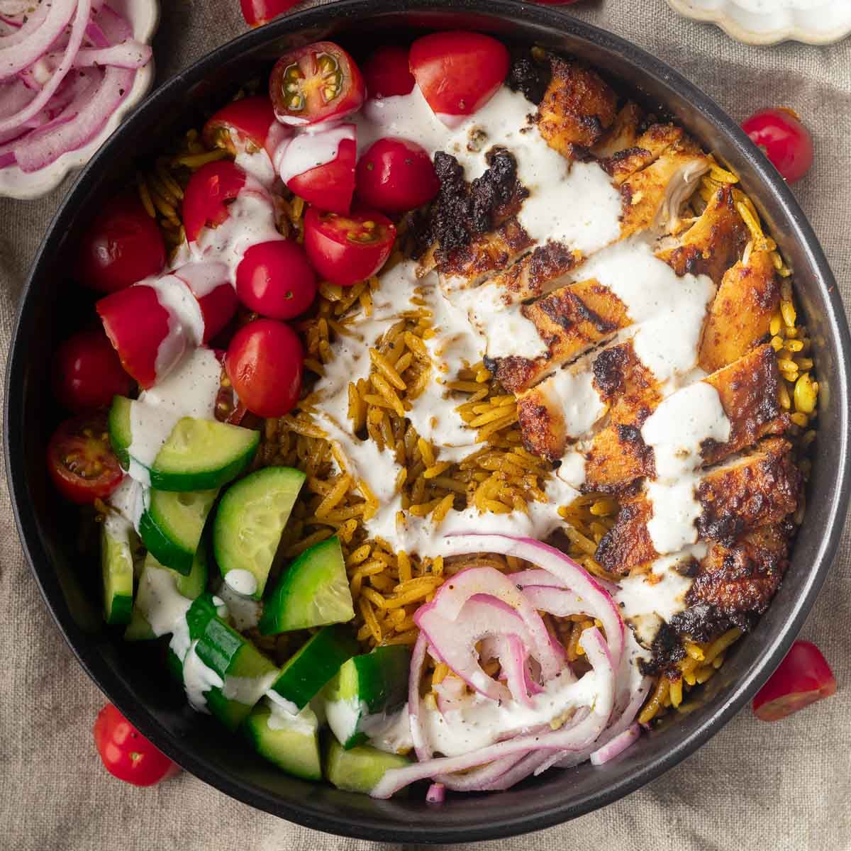 One pot shawarma and rice with grilled chicken bowl with rice, tomatoes, cucumbers, red onions, and white sauce on a black plate.