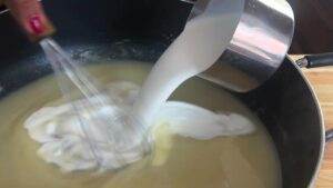 Pouring cream into saucepan, mixing ingredients for homemade soup or sauce preparation.