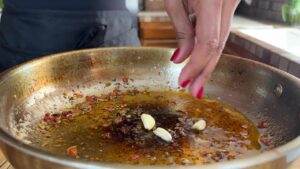 Hand adding garlic cloves to a sizzling pan with oil and spices, creating a flavorful base for cooking.