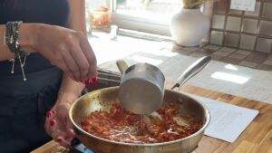 Woman adding liquid to tomato skillet sauce on a stove.