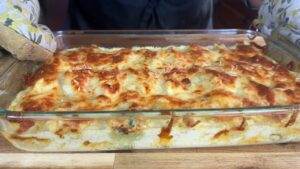 Golden cheesy casserole fresh from the oven on a wooden surface, wearing floral oven mitts.