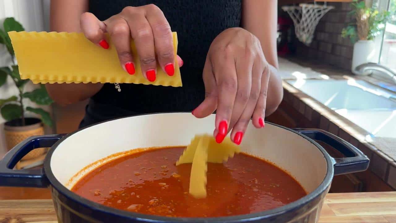 Hands adding lasagna noodles to a pot of tomato sauce on a wooden countertop in a kitchen setting.