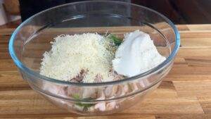 Glass bowl with shredded cheese, ricotta, herbs, and chicken on a wooden countertop, ready for mixing.