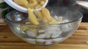 Cooling pasta shells in an ice water bath in a glass bowl on a wooden surface, emitting steam.