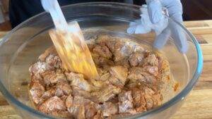 Person mixing marinated chicken in a glass bowl with a spatula on a wooden countertop.