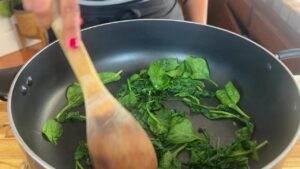 Stirring fresh spinach in a non-stick pan with a wooden spoon, close-up cooking scene in a kitchen.