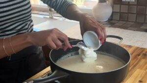 Person adding cream to a pan of cooking sauce in a kitchen.