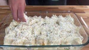 Hand adding cheese to creamy pasta bake in glass dish on wooden table.