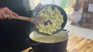 A person adding gnocchi to a pot of creamy soup on a wooden countertop.