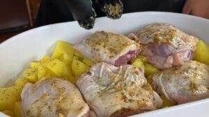 Seasoned chicken thighs on diced potatoes in a white baking dish, ready for the oven.