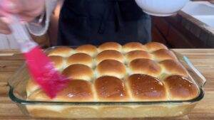 Freshly baked dinner rolls being brushed with butter in a glass baking dish on a wooden countertop.