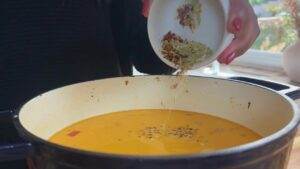 Person adding spices to a pot of creamy soup, enhancing flavor and aroma in a cozy kitchen setting.