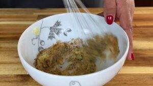 Hand whisking spices in a white ceramic bowl on a wooden surface.