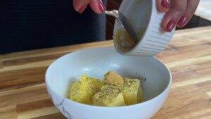 Hand adding butter to seasoned potatoes in a bowl on a wooden surface, highlighting a cooking preparation process.