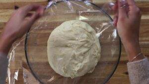 Person covers dough in a glass bowl with plastic wrap, preparing it to rise.