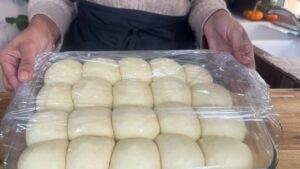 Person holding a tray of dough balls covered in plastic wrap, ready to rise in a kitchen setting.