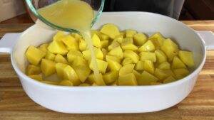 Pouring broth over diced potatoes in a baking dish on a wooden countertop.