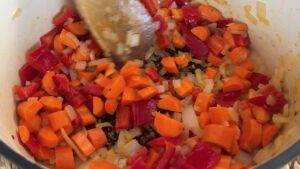 Chopped carrots, red bell peppers, and onions being sautéed in a pot, stirred with a wooden spoon.