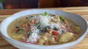 Creamy gnocchi soup topped with grated cheese and fresh herbs in a white bowl on a wooden table.