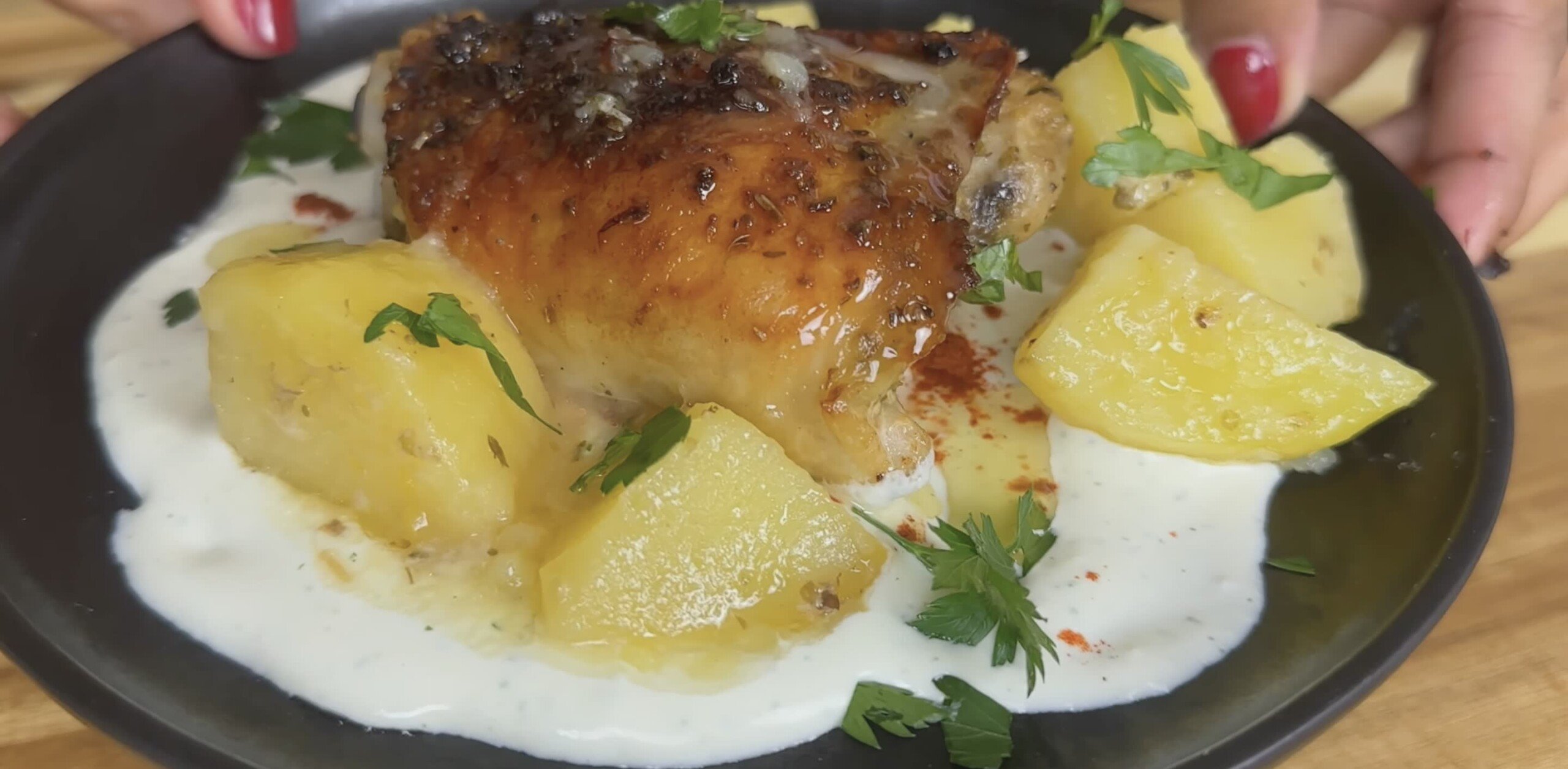 Roasted chicken with golden potatoes on creamy sauce, garnished with fresh parsley on a black plate.