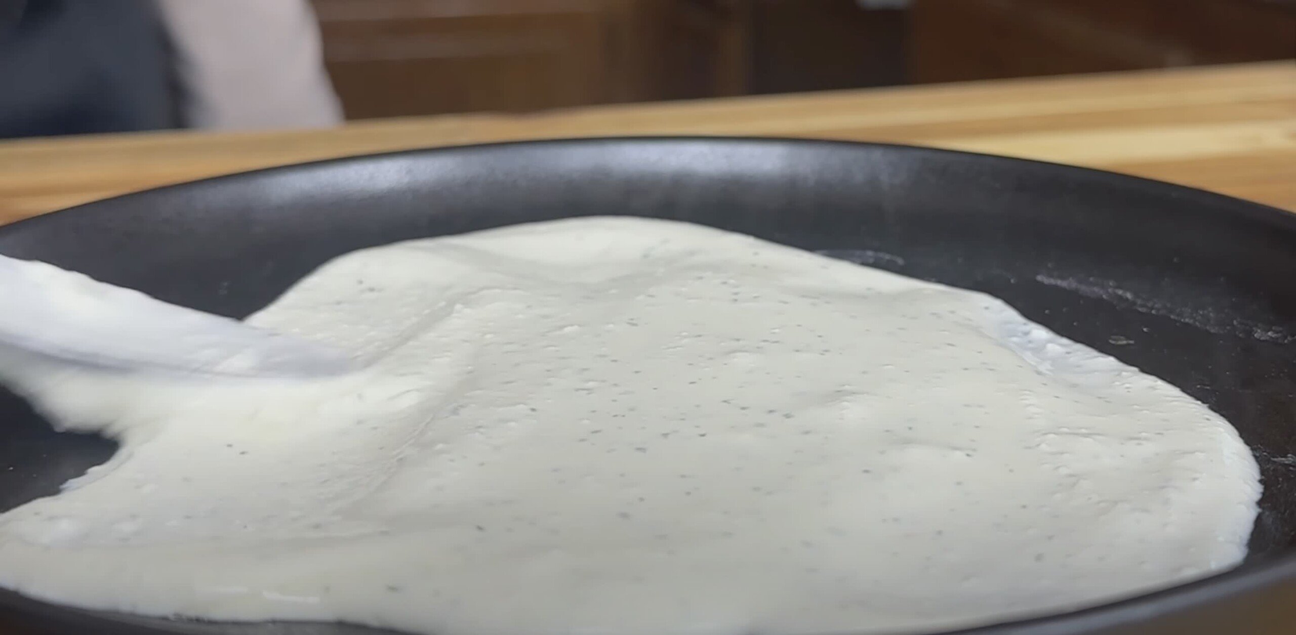 Pancake batter being spread on a black skillet with a spatula in a kitchen setting.