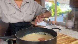 Person adding spice to creamy sauce in a pan in kitchen with sunlight from window.