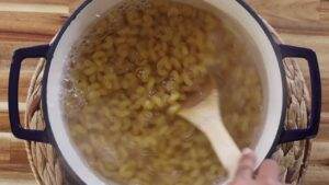 Boiling elbow pasta in a pot with a wooden spoon stirring, on a wooden table.