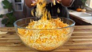 Hands adding shredded cheese to a glass bowl on a wooden countertop.