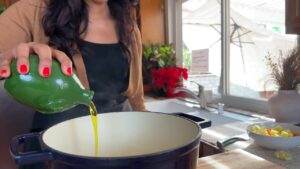 Woman pouring olive oil into a pot in a sunny kitchen with fresh ingredients ready for cooking.