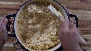 Person stirring creamy homemade mac and cheese in a pot on a wooden table.