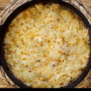 Cheesy potato gratin in a black skillet, topped with golden-brown cheese, displayed on a woven mat.