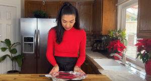 Woman in red sweater prepares food in cozy kitchen with poinsettia plants.