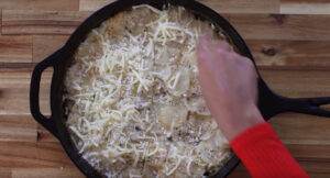 Hand adding cheese to creamy potato gratin in a cast iron skillet on wooden surface.