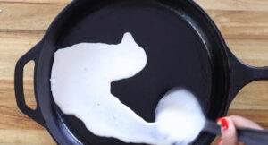 Pouring dosa batter onto a hot skillet for cooking, on a wooden table background.