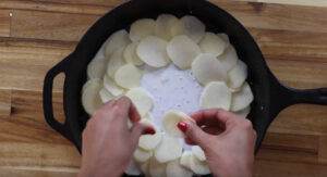 Hands arranging sliced potatoes in a cast iron skillet with cream, preparing potato gratin on a wooden table.