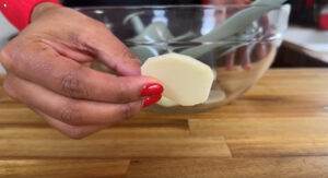 Hand holding a thinly sliced potato above a glass bowl on a wooden counter, close-up.