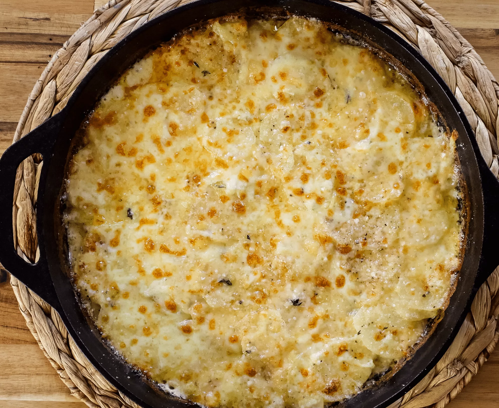 Cheesy potato gratin in a black skillet, topped with golden-brown cheese, displayed on a woven mat.