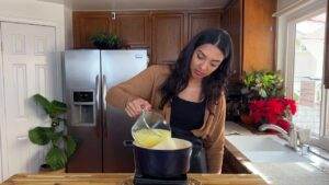 Woman in kitchen pouring mixture into pot, surrounded by plants and flowers, near stainless steel fridge.