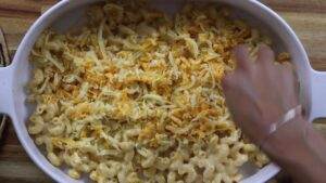 Cheesy baked mac and cheese being prepared in a white casserole dish on a wooden table.