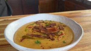 Creamy soup topped with crispy garnish and herbs in a white bowl on a wooden table.