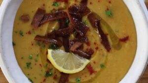 A bowl of creamy lentil soup garnished with lemon slice, herbs, crispy croutons, and spices.