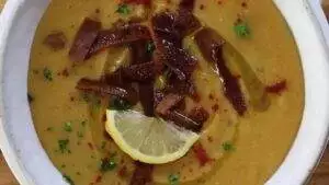 A bowl of creamy lentil soup garnished with lemon slice, herbs, crispy croutons, and spices.