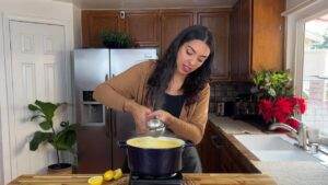 Woman in kitchen squeezing lemon juice into a pot on the stove, surrounded by fresh ingredients and plants.