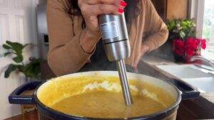 Woman blending homemade soup in a pot with an immersion blender in a cozy kitchen setup.