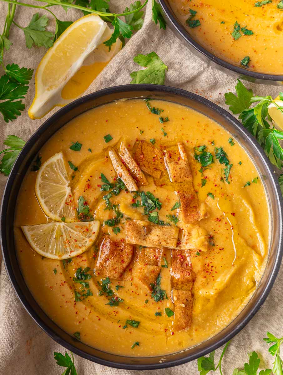 Bowl of Lebanese lentil soup with lemon slices, pita croutons, and parsley garnish on beige cloth.