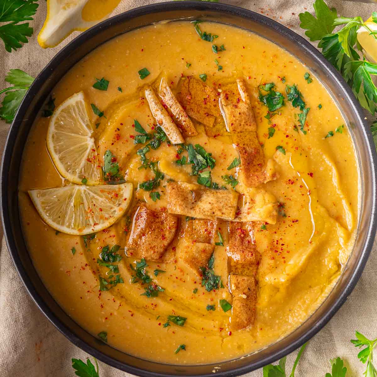 Bowl of Lebanese lentil soup topped with lemon slices, crispy bread strips, parsley, and red pepper flakes, garnished with herbs.