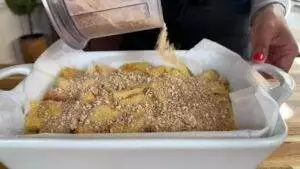 Sprinkling crunch topping on banana dessert in a white baking dish on a wooden table.