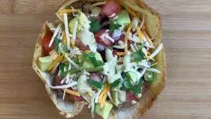 Taco salad in a crispy shell topped with avocado, cheese, tomatoes, cilantro, and creamy dressing on a wooden board.