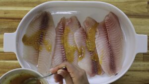 Marinating fish fillets with seasoned dressing in a white dish on a wooden table.